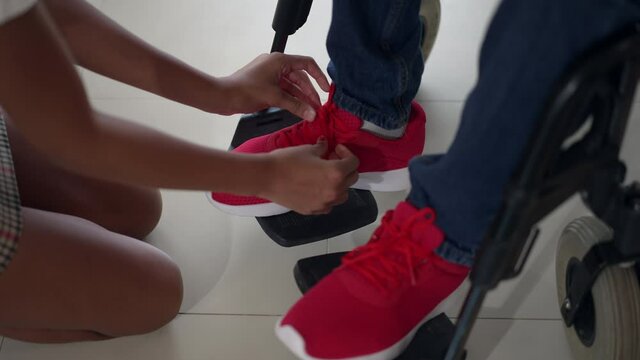 High angle view feet of disabled man on legrest with unrecognizable teenage girl tying shoelaces on red sneakers. African American adolescent daughter helping ill father indoors at home