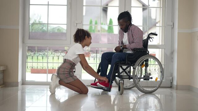 Wide shot positive teenage African American daughter taking care of disabled father tying shoelaces. Side view portrait smiling girl and man in wheelchair talking putting on shoes at home indoors