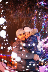 young man hold little girl in arms on town square decorated with Christmas lights, garlands,...