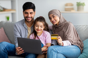 Loving arabic family of three with laptop and credit card making online shopping, sitting together on sofa