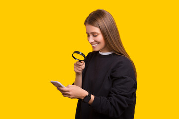 Portrait of cute girl looking at mobile phone through magnifying glass over yellow background