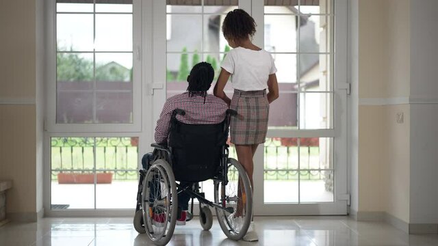 Back view disabled father and teenage daughter looking out the window talking. Wide shot African American man in wheelchair and girl hugging parent standing in living room at home. Family concept