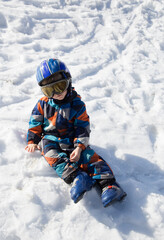 Fototapeta na wymiar tired kid in a helmet, glasses, ski boots and winter overalls sits in the snow at a ski resort. Children's skiing lesson at an alpine school. Fell in the snow, pause to rest. Winter activities