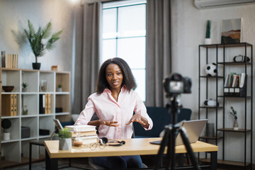 Charming african american woman in pink shirt talking and gesturing while recording video on modern camera. Female blogger sitting at home and doing live stream.