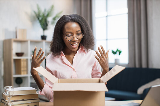 Attractive african woman with surprised facial expression looking inside paper box. Happy young lady in eyeglasses and pink shirt unpacking parcel.