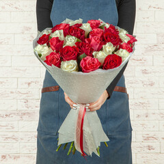 Young girl florist in a blue apron holds a fresh bouquet of red flowers wrapped in paper with a...