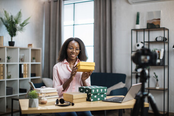 Pretty african woman in eyeglasses and casual wear recording video on camera while unpacking gift boxes. Female blogger sharing her emotions with her subscribers in social networks.