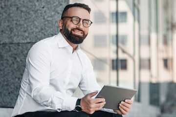 Fototapeta premium Confident middle-aged businessman working on tablet outdoors, sitting near modern office center, copy space