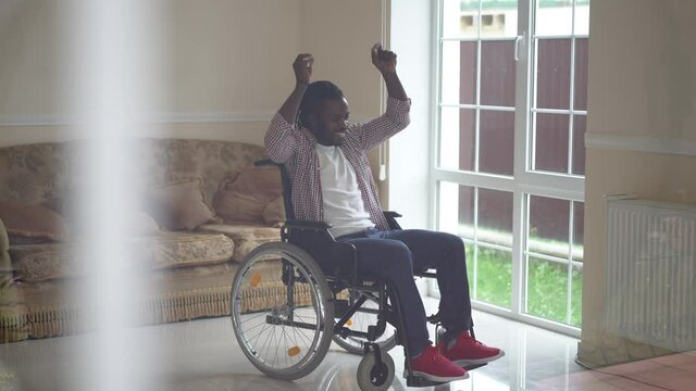 Happy African American disabled man rejoicing in slow motion dancing in wheelchair indoors. Wide shot portrait smiling handsome paralyzed guy gesturing success at home in living room