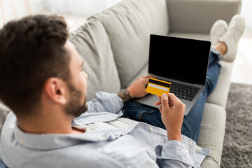 Shopaholism and e-commerce. Young guy holding credit card, shopping online via laptop, sitting on sofa at home