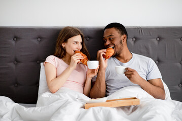 Obraz na płótnie Canvas Affectionate multiracial couple having breakfast in bed, eating fresh croissants wth coffee at home