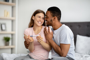 Excited multinational couple with positive pregnancy test hugging on bed, celebrating successful...