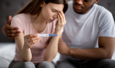 Upset young woman showing negative pregnancy test, her black husband supporting her in bedroom, selective focus