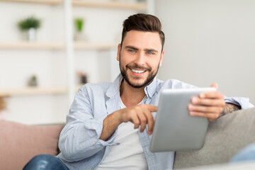 Handsome young man holding digital tablet and smiling at camera, sitting on couch at home, free space