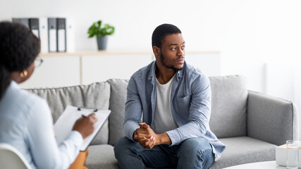 Depressed black man consulting psychologist, sitting on couch deep in thought, panorama