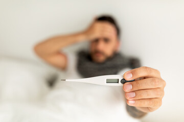 Sick young man showing thermometer to camera, lying on bed and touching forehead. Selective focus, top view