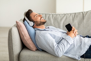 Fototapeta premium Happy relaxed guy in wireless headset lying on sofa with closed eyes and smiling while listening to music