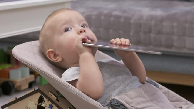 9-month-old baby boy using a digital tablet computer as baby teether toy, cute little child holding mobile device sitting on rocking chair. Children and mobile technologies. High quality 4k footage