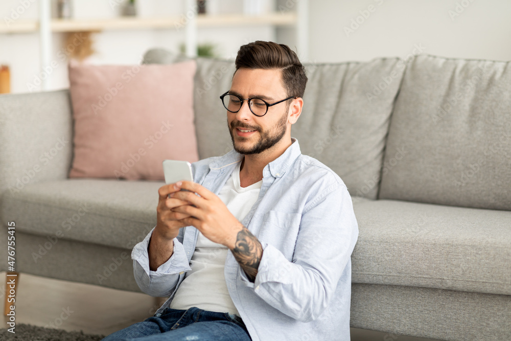 Poster useful app. handsome millennial man using smatphone while resting at home, sitting on floor and lean