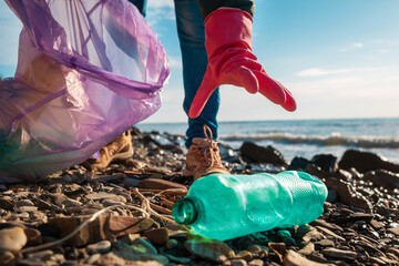 A volunteer in rubber gloves reaches for a dirty plastic bottle lying on the ocean shore. Hand...