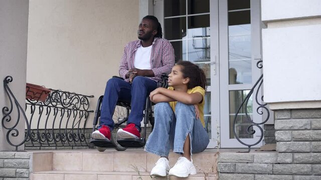 Wide shot portrait relaxed carefree African American daughter and disabled father on porch looking away talking. Happy confident teenage girl and man enjoying tranquility on backyard or front yard