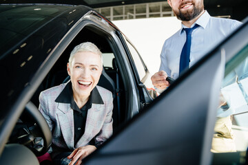 Gorgeous blond woman who is sat in car in car dealership smiling while receiving car keys to her...