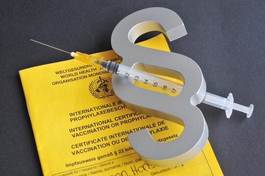 Hamburg, Germany - December 22, 2021: Close up of a hypodermic syringe, a paragraph sign and Vaccination book in the background - Symbol for the legal aspects of mandatory vaccination