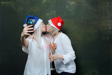two girlfriends kissing on the lips in bright  santa hats, having fun drinking champagne and taking...