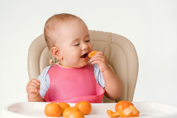 A baby in a pink bib sits on a chair and eats tangerines with an appetite
