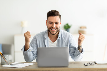 Joyful young man shaking fists looking at laptop screen celebrating business success, sitting at workplace at home
