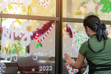 Asian woman decorates beautiful stickers on window glass in front of cafe to welcome Christmas and New Year. Prepare to decorate the storefront to be beautiful during the important holidays of year.