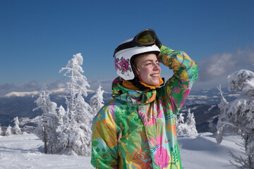 Fototapeta na wymiar satisfied teenager girl in a ski suit, helmet, stands in the mountains against the background of icy trees on a sunny winter day. a minute of rest, relaxation, enjoyment, winter holidays