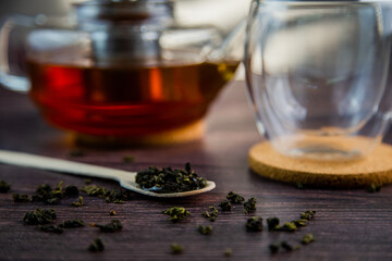 Fototapeta premium A wooden spoon with tea lies against a dark wood background, a mug and a teapot are visible from behind, a photo in dark colors. High quality photo