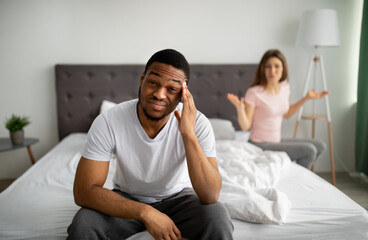 Displeased young guy sitting on bed, massaging temples in irritation, his angry wife shouting at...