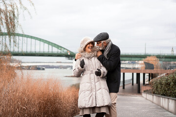 An elderly couple is walking around the waterfront in an urban area. The couple is smiling and looking happy in love.