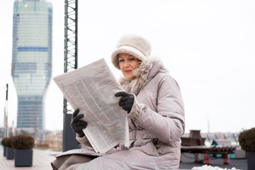 A beautiful, elegant senior woman sits by the river and reads the newspaper on a cold winter day.  She is wearing a long winter jacket with fur around the neck and a warm hat and gloves.