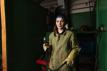 Young welder in uniform holding welding torch and looking at camera in factory