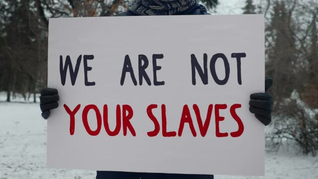 Person is standing in the Cold and holding a White Sign that says We Are Not Your Slaves. Man taking Part in a Protest Against the Government Policies that are Unfair to People of Color. 