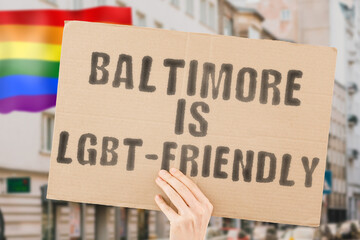 The phrase " Baltimore is LGBT-Friendly " on a banner in men's hand with blurred LGBT flag on the background. Human relationships. different. Diverse. liberty. Sexuality. Social issues. Society