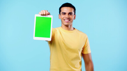 pleased young man in yellow t-shirt holding digital tablet with green screen isolated on blue.