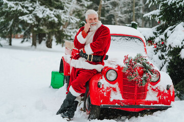 fashionable Santa Claus with a gray beard stands in the woods near a red pickup truck with gifts. High quality photo - obrazy, fototapety, plakaty