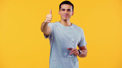 happy young man in t-shirt holding bottle with water and showing thumb up isolated on yellow.