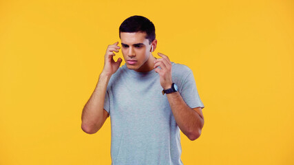 confused young man in t-shirt adjusting wireless earphones isolated on yellow.