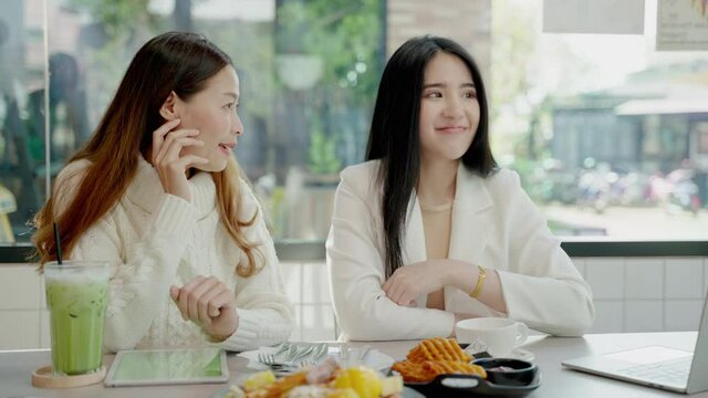 Two beautiful Asian female friends are sitting happily eating and chatting about the taste of food in a cafe, urban lifestyle concept, vacation relaxation.

