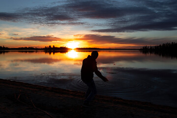 silhouette of Young Filipino man skipping rocks on lake during colourful sunset