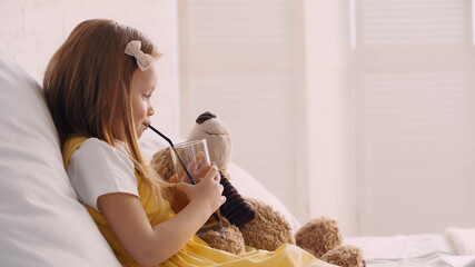 Side view of kid drinking juice near teddy bear on bed.