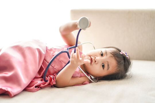Asian cute little girl in a beautiful pink dress holding a stethoscope and role-playing as a doctor, lying down on sofa in the living room.