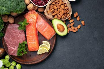 Green vegetables, nuts, beans with beef meat and salmon fish on dark background with copy space top view. Raw healthy food, clean eating or balanced diet concept