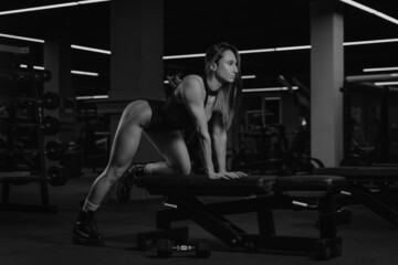 Fototapeta na wymiar A fit brunette woman is stretching after a single-arm dumbbell row with her knee on the bench. A muscular girl wears a black stringbody and boots during a back workout in a gym.