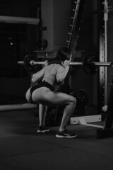 A sporty woman with dark hair is squatting with a barbell near the squat rack in a gym. A girl in shorts is doing a leg workout.